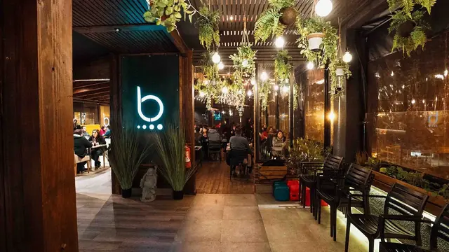 Entrada de B-Restobar con su letrero iluminado, plantas colgantes y mesas al fondo
